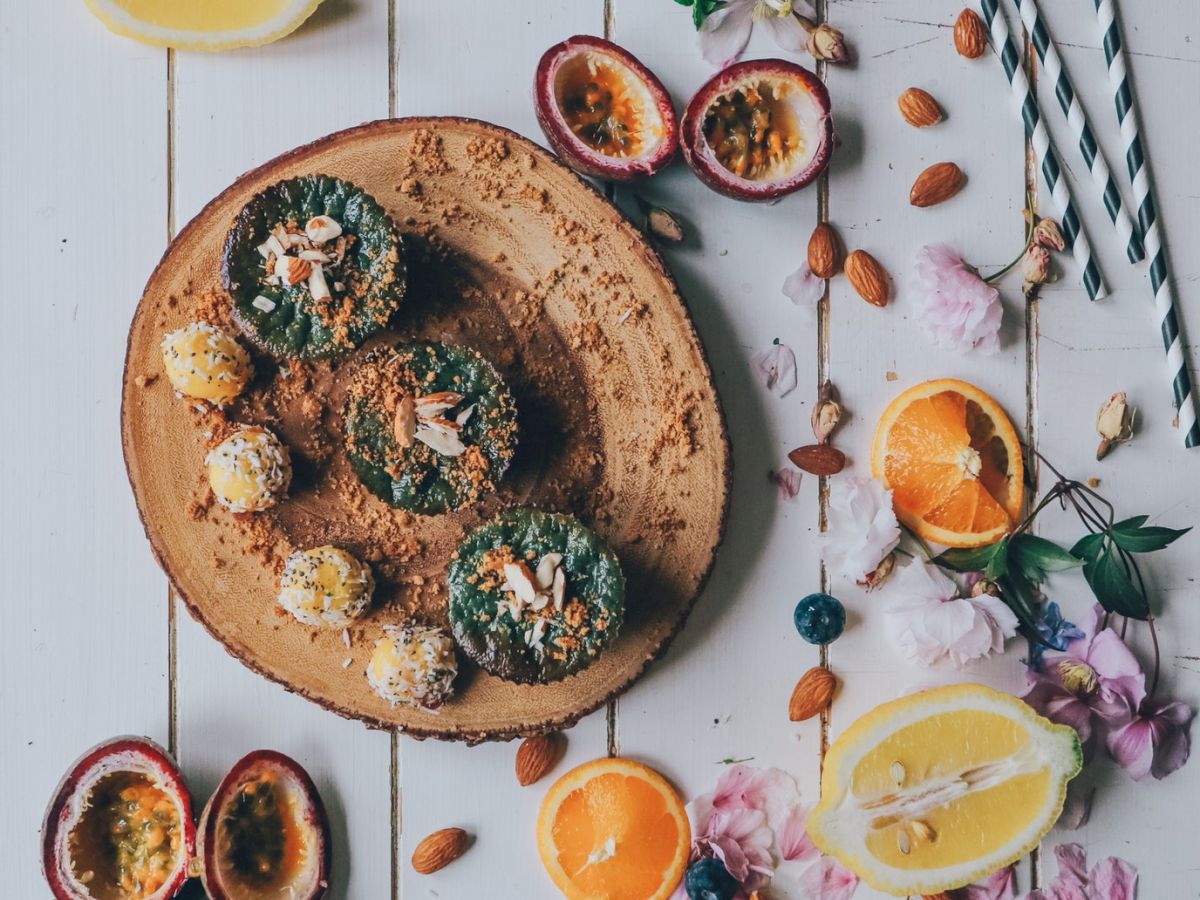 The image shows a wooden platter with baked goods surrounded by halved oranges, passion fruits, almonds, lemon slices, flowers, and striped straws.