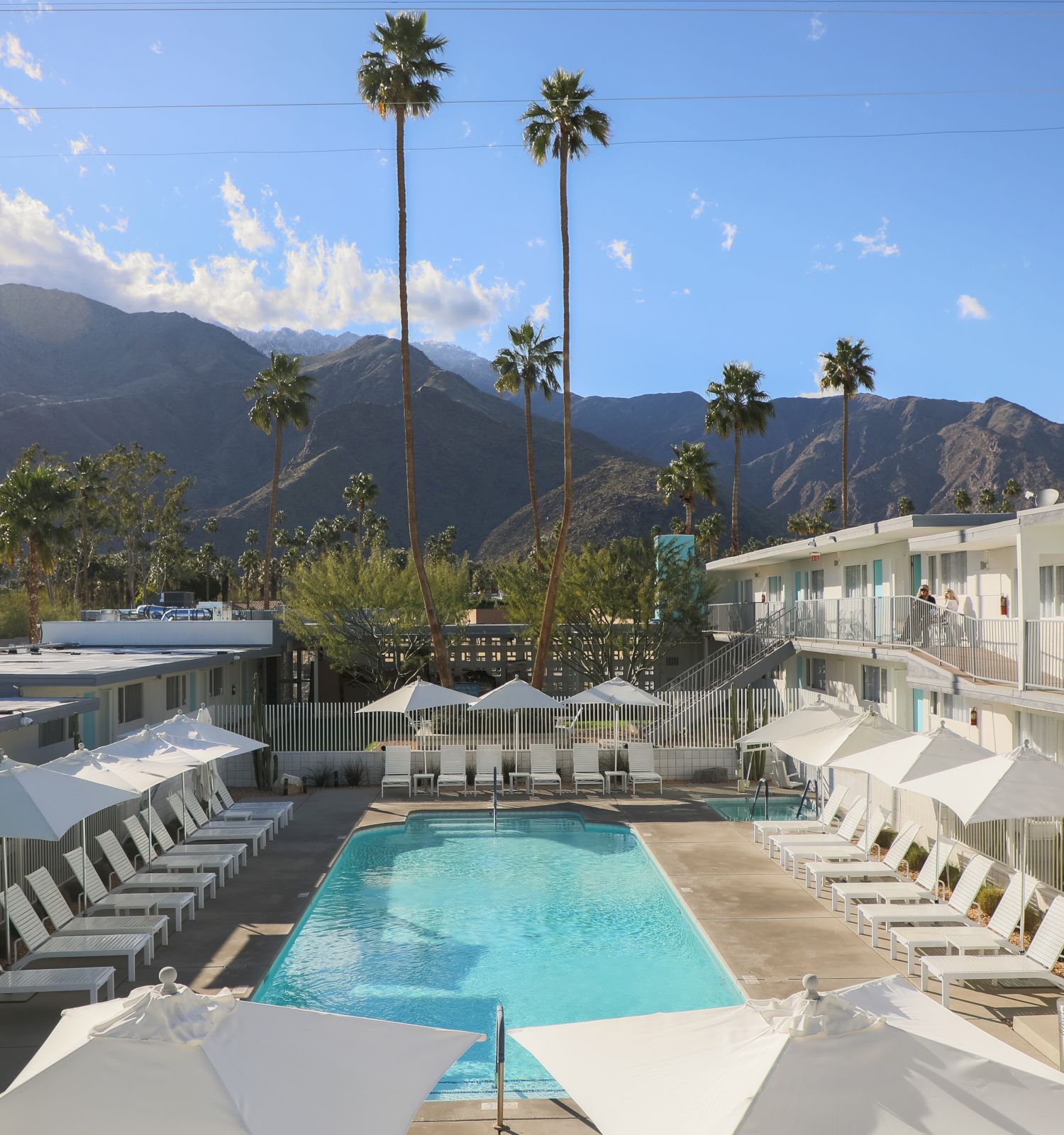 A serene hotel pool area with lounge chairs, umbrellas, and a mountainous backdrop under a clear sky with palm trees scattered around the site.