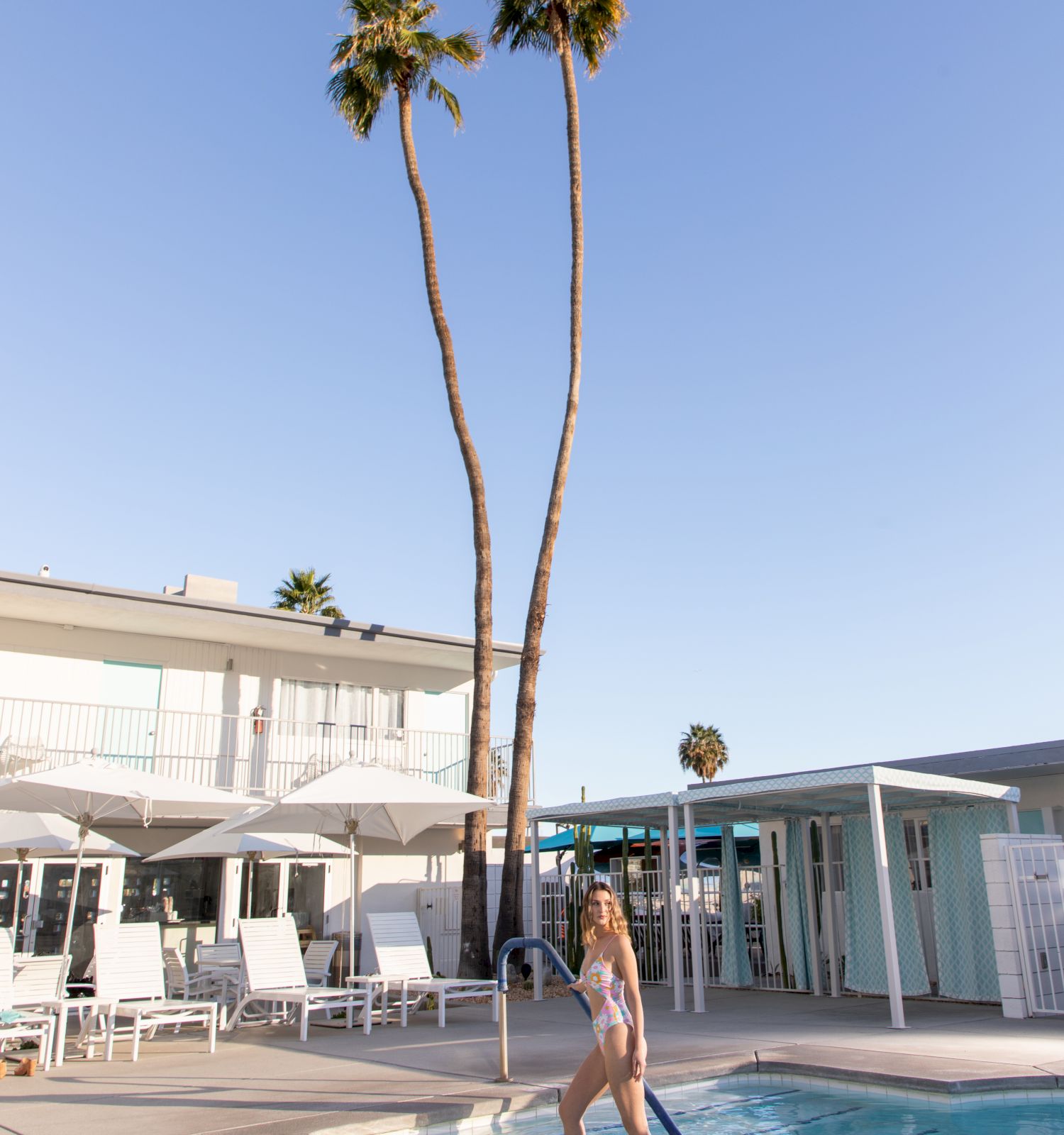 A person in swimwear wades into a pool near a modern building with white loungers and umbrellas, and tall palm trees in the background under a clear sky.