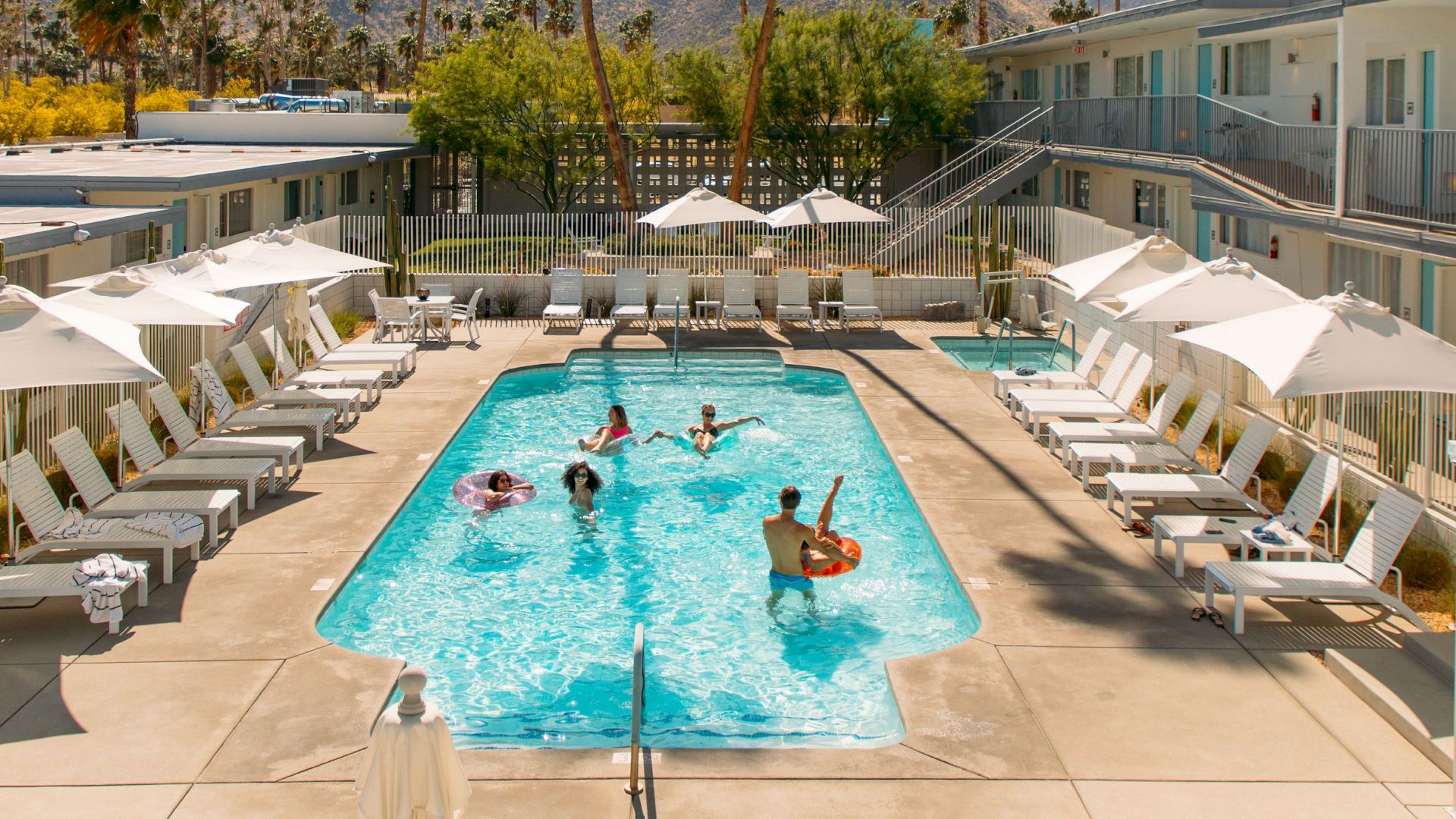 A pool area at a resort or motel with people swimming, surrounded by lounge chairs, umbrellas, and two-story buildings in the background.