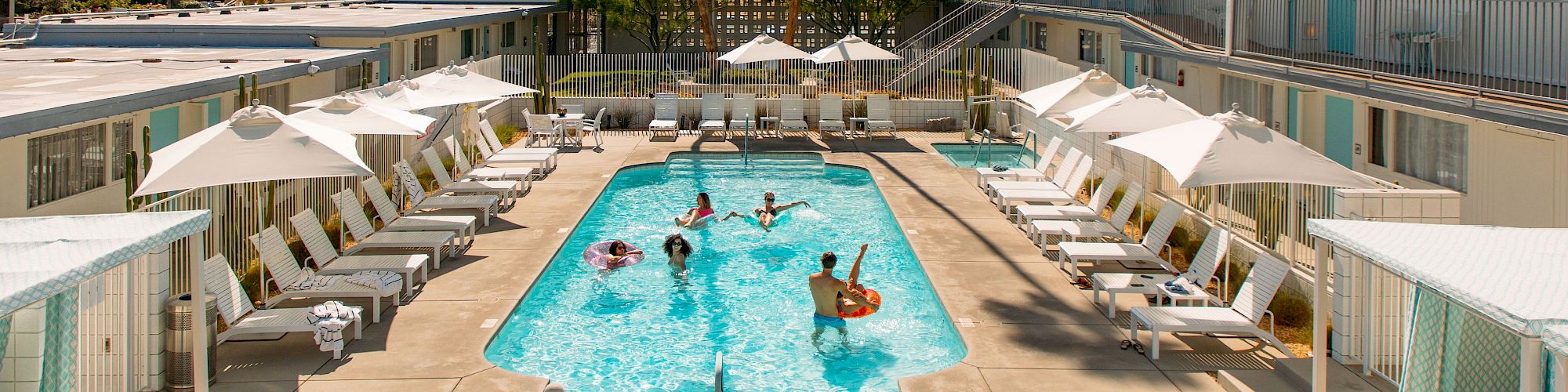 A pool area at a resort or motel with people swimming, surrounded by lounge chairs, umbrellas, and two-story buildings in the background.