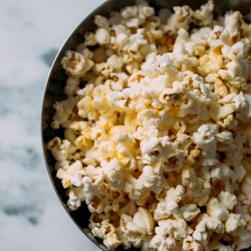 A bowl filled with freshly popped popcorn is placed on a marble surface. The popcorn appears fluffy and lightly buttered, ready to be enjoyed.