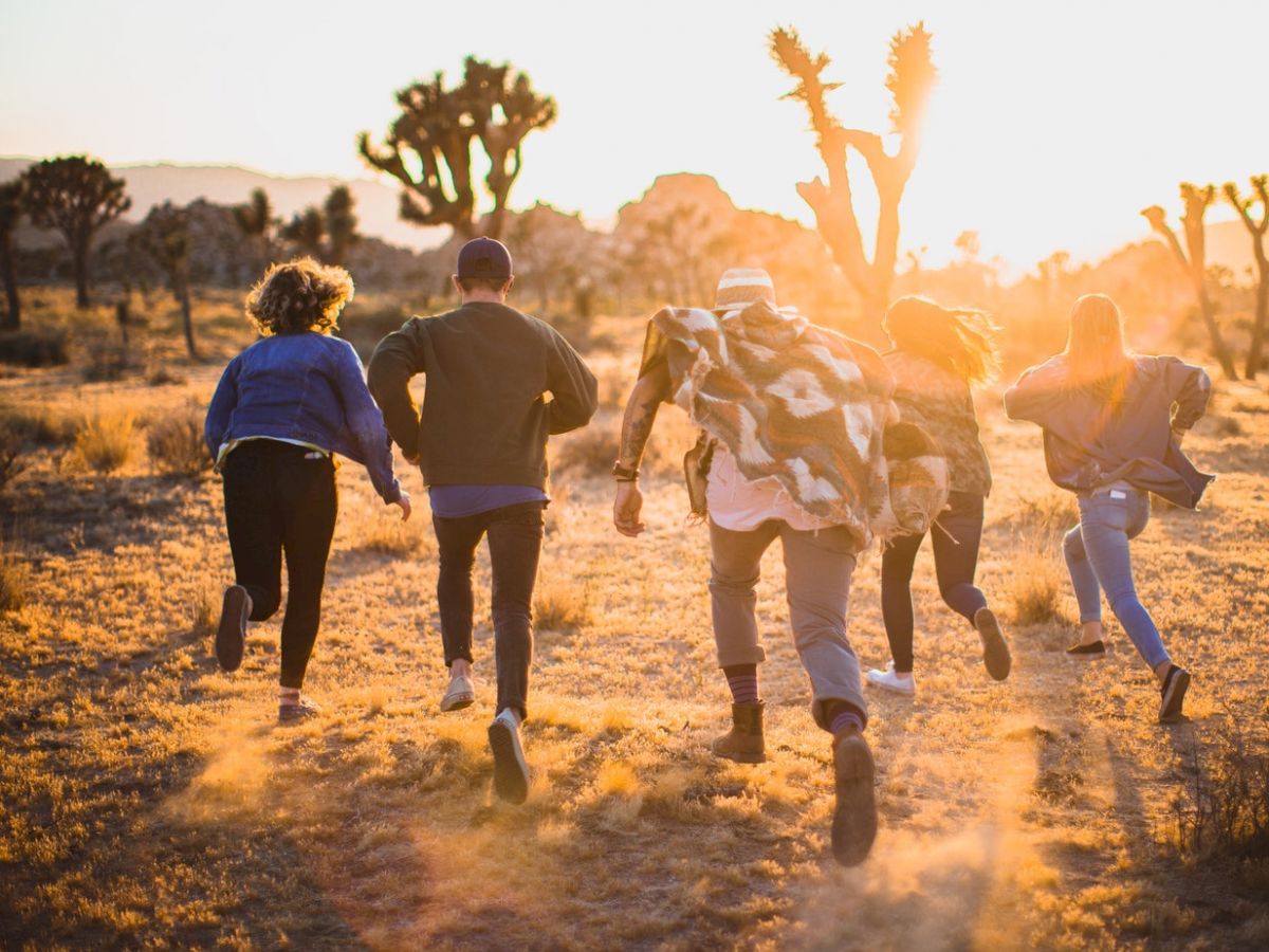 A group of five people running through a desert landscape during sunset with dust kicking up from their steps, Joshua trees in the background.