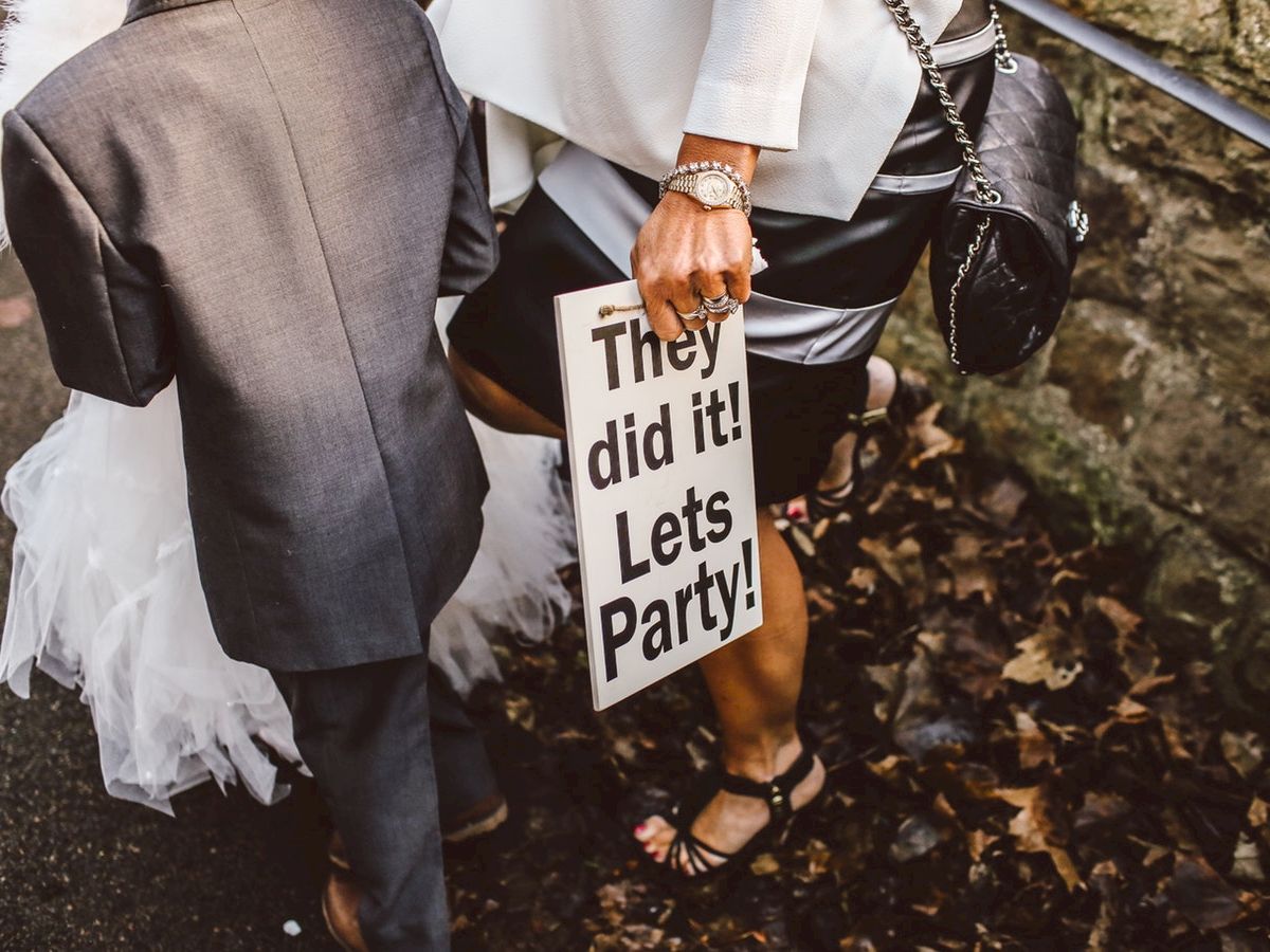 A person holds a sign reading "They did it! Let's Party!" while walking with a child dressed in a suit along a path with fallen leaves.