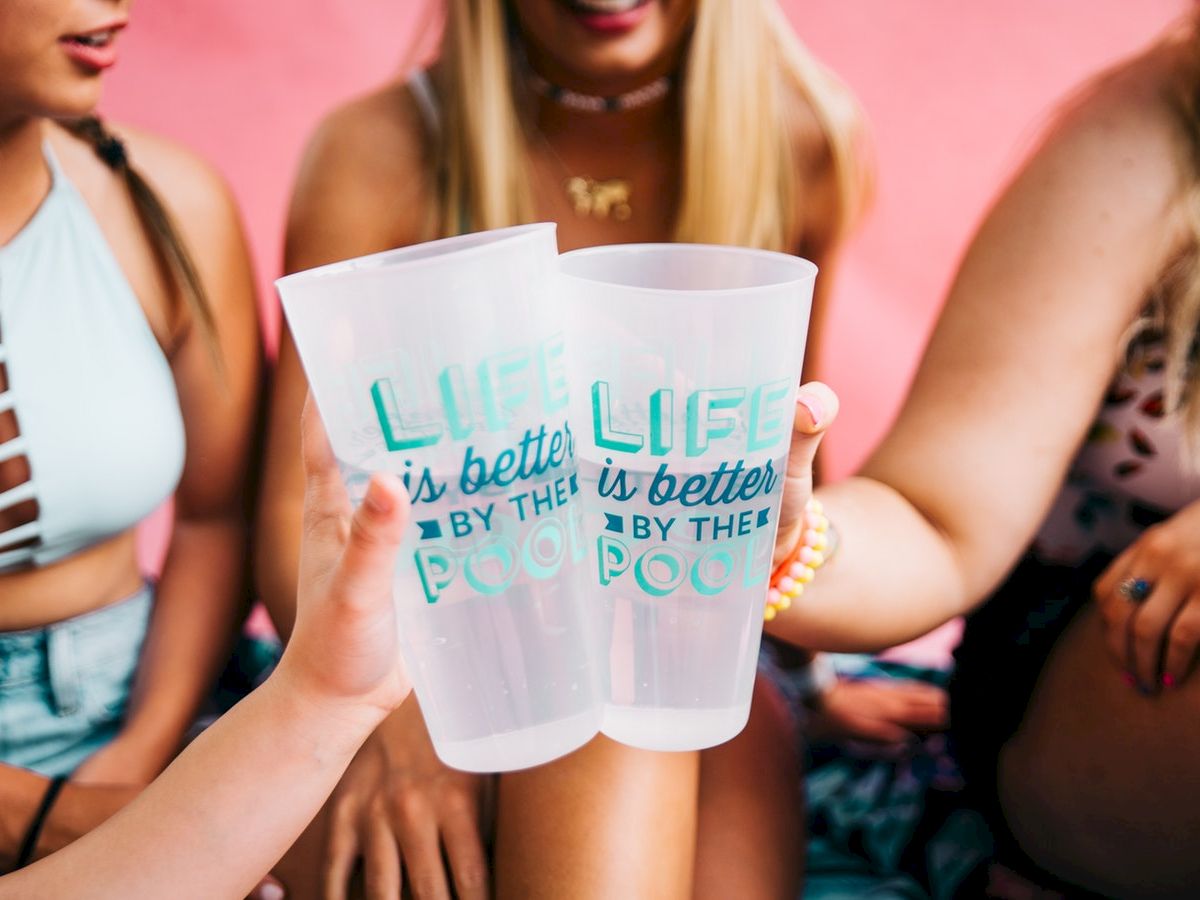 Three people are holding and clinking plastic cups that read "LIFE is better BY THE POOL." They are sitting together.