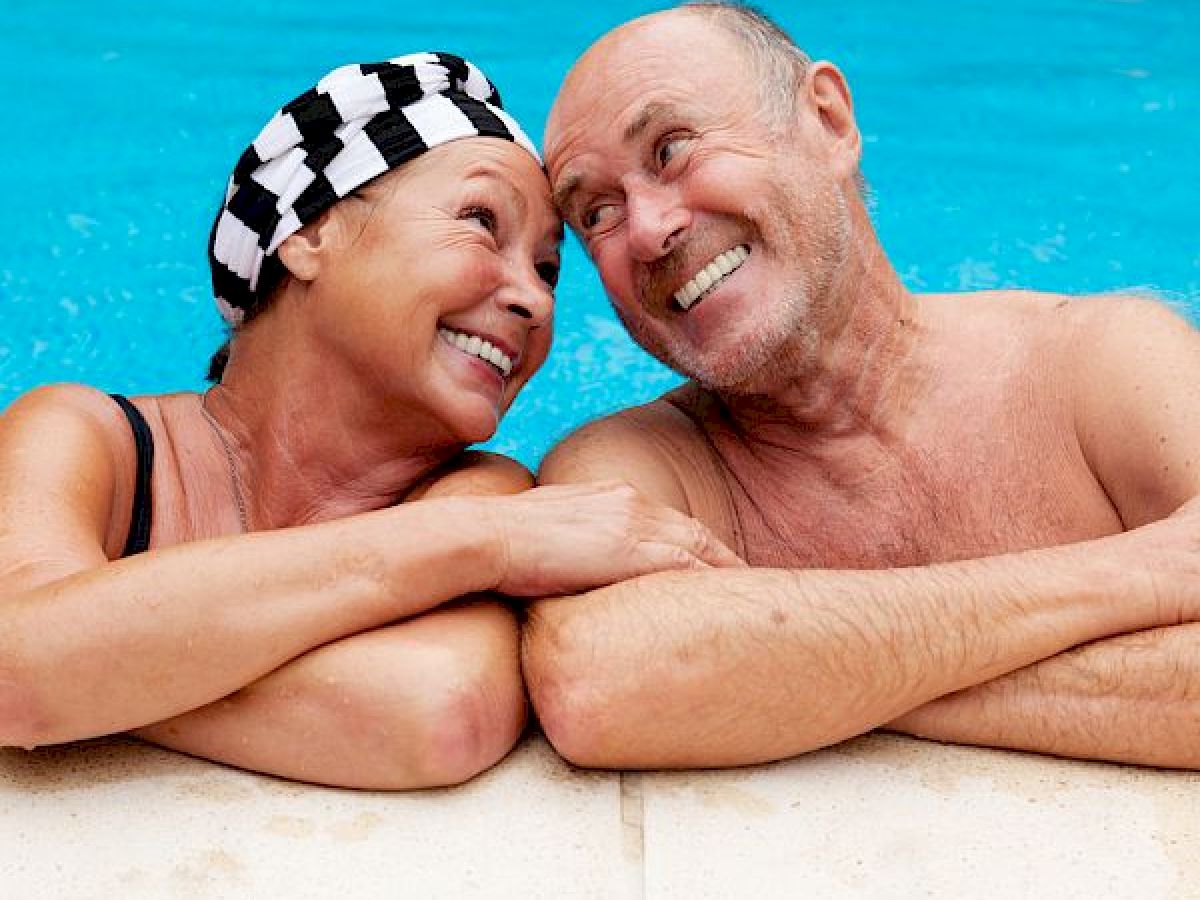 Two smiling older adults, a woman and a man, are relaxing at the edge of a swimming pool with their arms resting on the poolside.