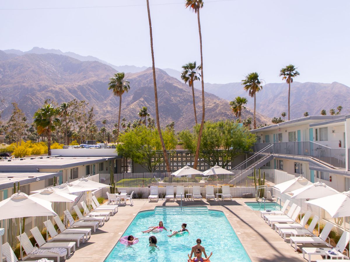 A serene outdoor pool area with people swimming, surrounded by lounge chairs and umbrellas, set against a backdrop of palm trees and mountains.