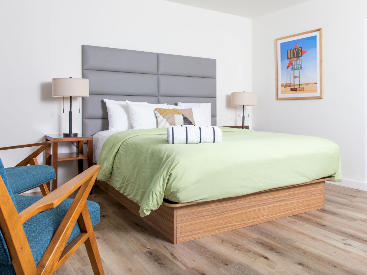 A modern hotel room with a large bed, green bedding, wooden furniture, two bedside tables with lamps, and a framed picture on the wall.