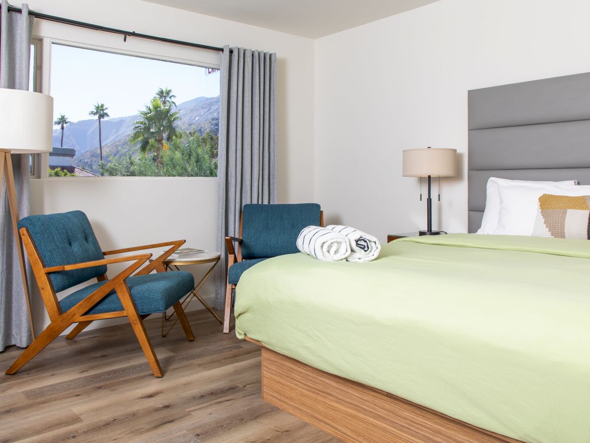 A modern bedroom with wooden flooring, a large bed, armchairs, a floor lamp, and a window with a view of palm trees and mountains outside the window.