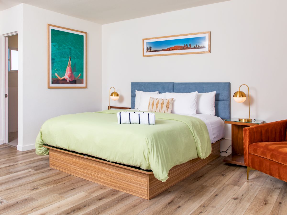 A modern bedroom with a green bedspread, orange chair, wall art, side tables, lamps, and a large mirror on the left wall.