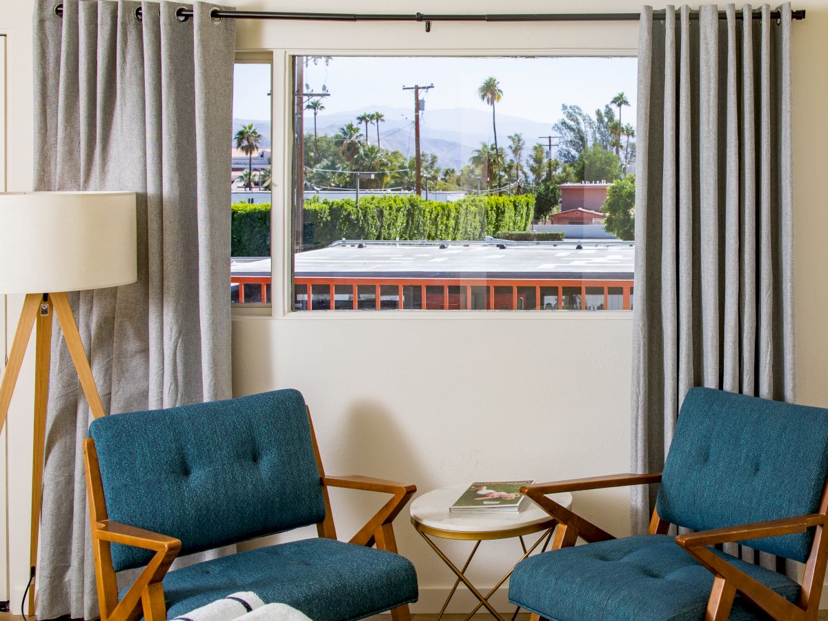 Two blue chairs and a small round table are in front of a window with gray curtains, offering an outside view of palm trees and buildings.