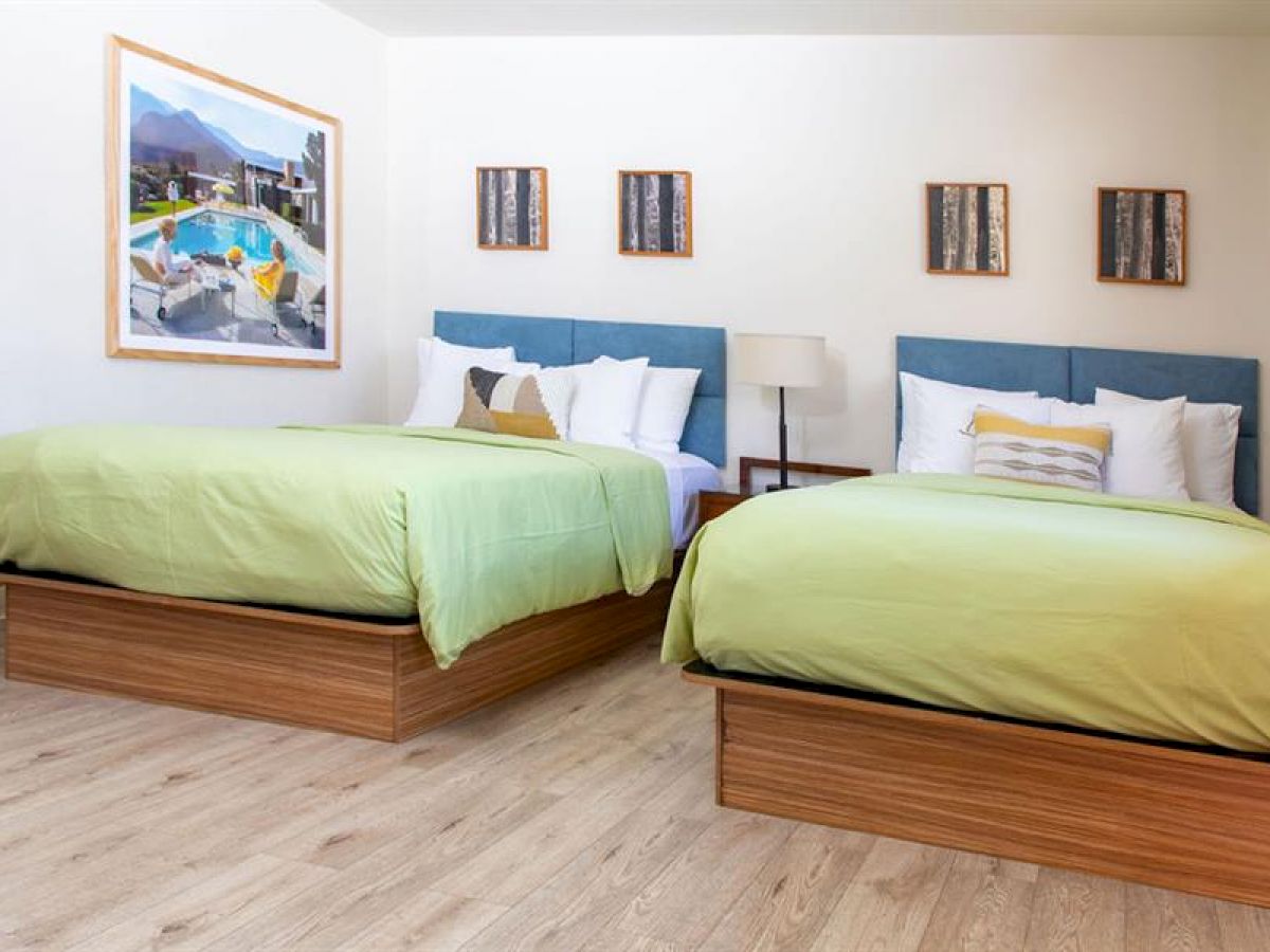 The image shows a neatly arranged hotel room with two green double beds, a painting on the wall, three small frames, and a bedside lamp.