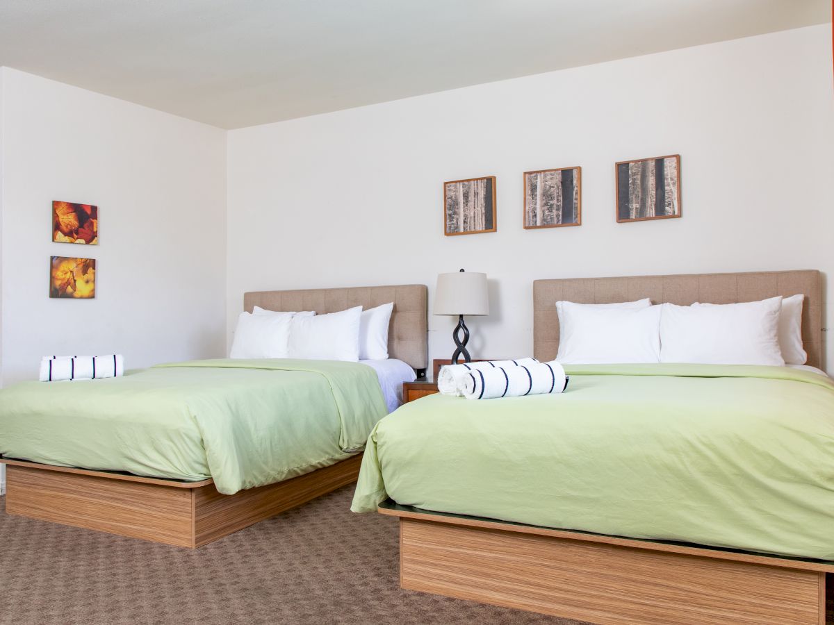 A hotel room with two double beds, green bedding, white pillows, rolled towels, wall art, an orange curtain, carpeted floor, and a bedside lamp.