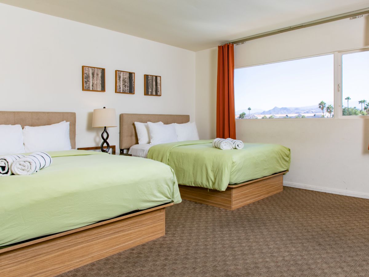 This image shows a hotel room with two double beds, green bedding, rolled towels, a side table with a lamp, and a window with a scenic view ending the sentence.