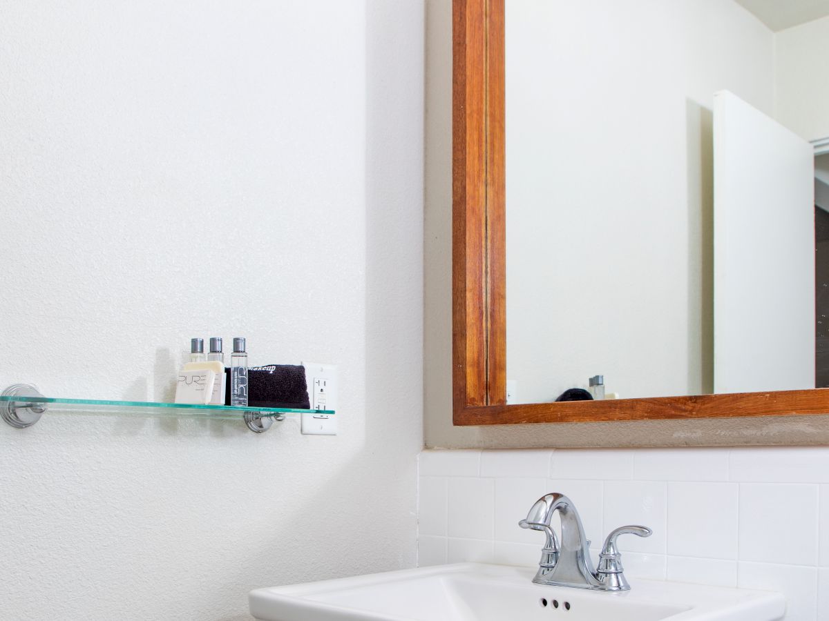 The image shows a bathroom with a sink, a large mirror with a wooden frame, and a glass shelf holding toiletries.