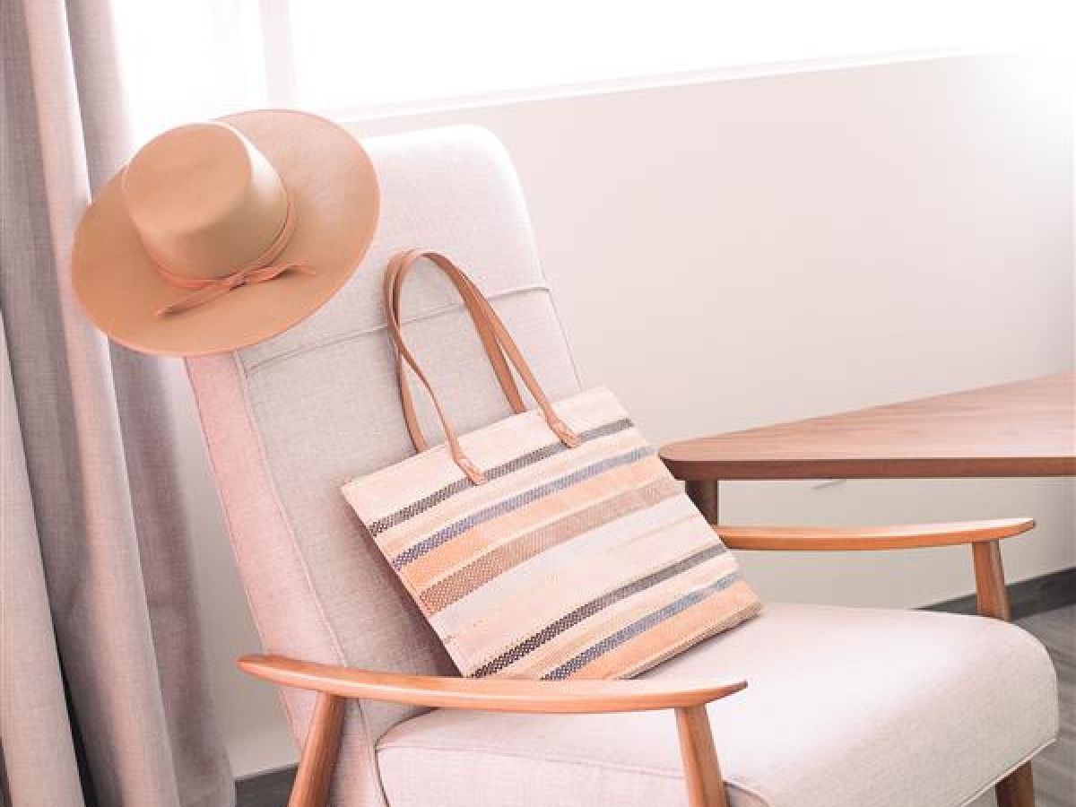 A cozy chair with wooden armrests, holding a wide-brimmed hat and a striped tote bag, positioned next to a table and curtains.