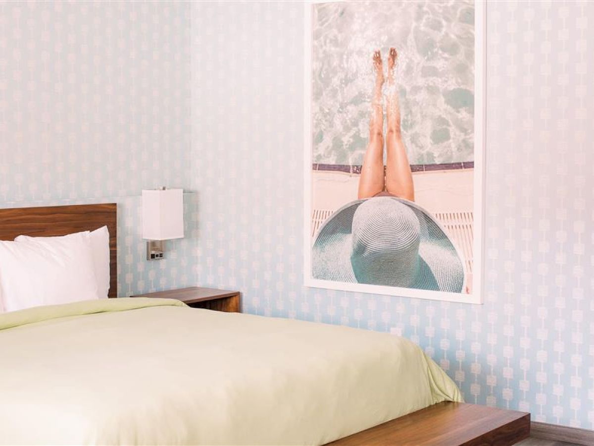 A neatly-made bed with a green blanket, wooden headboard, wall-mounted lamp, and a wall art showing a person lounging by a pool.