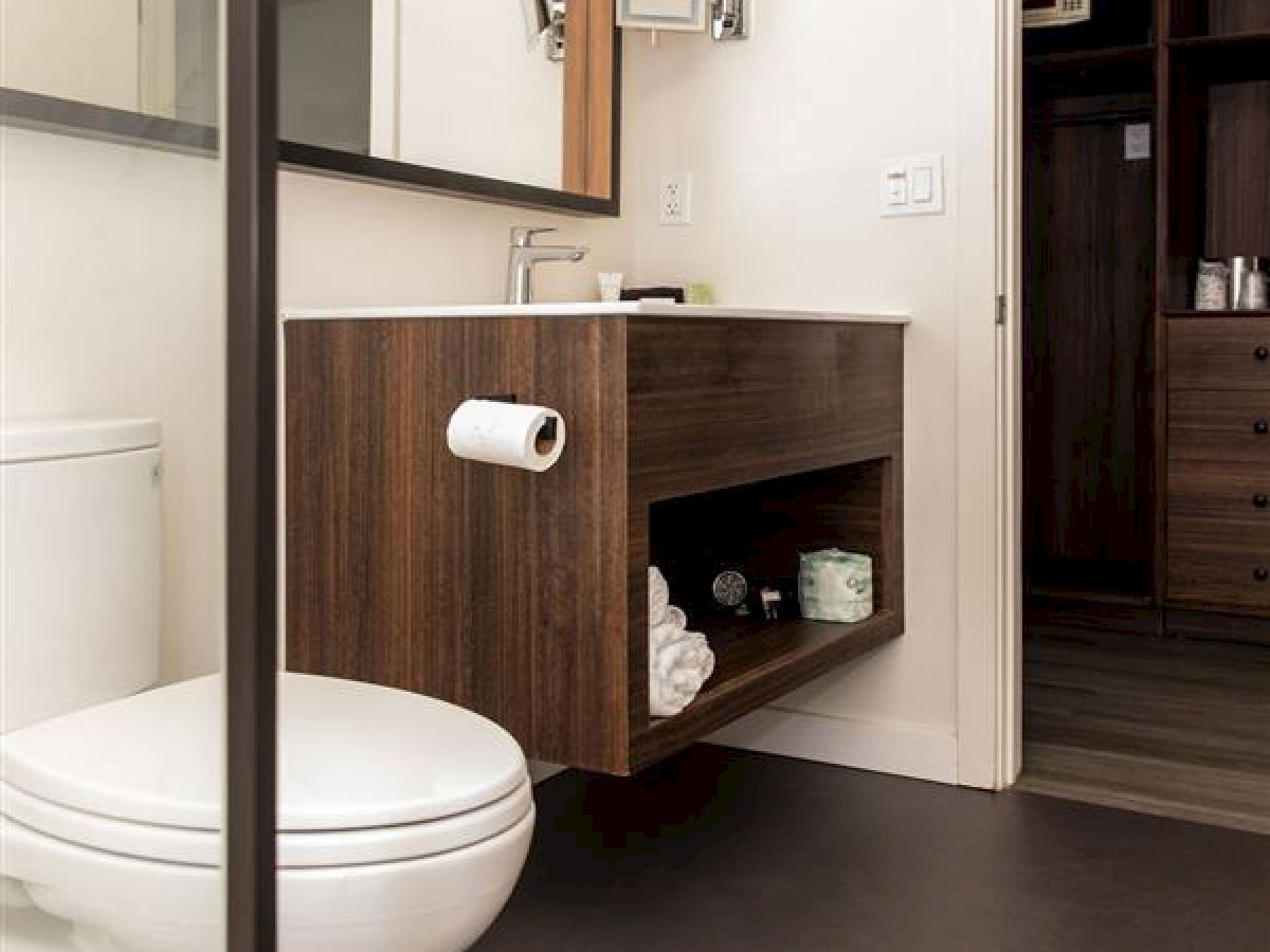 The image shows a modern bathroom with a toilet, floating wooden vanity, large mirror, and an open door leading to another room.