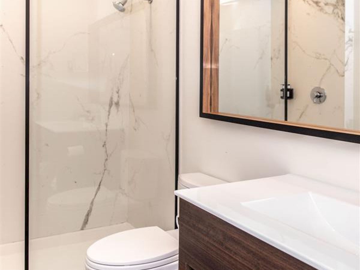 The image shows a modern bathroom with a shower, toilet, large mirror, and a wooden vanity with a white countertop and open shelves holding towels.