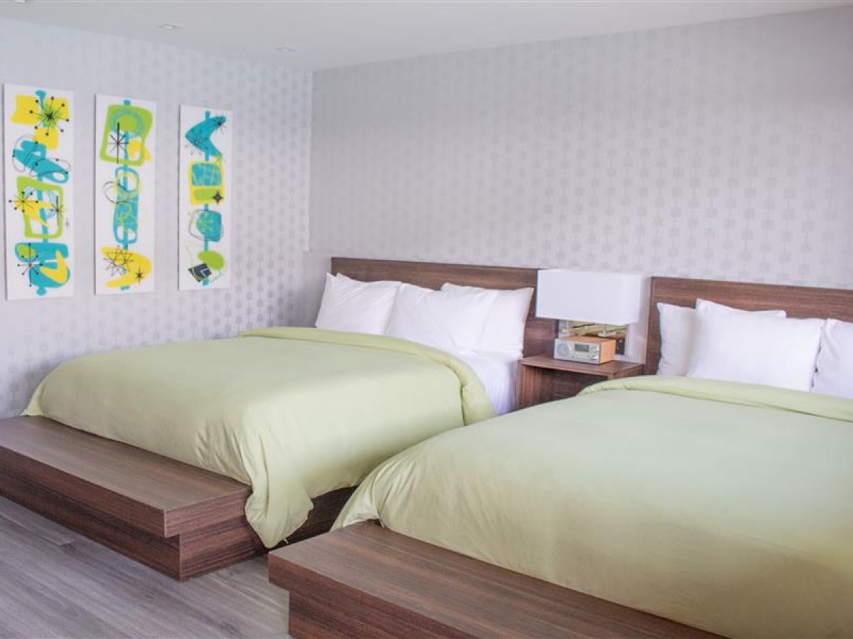 The image shows a tidy room with two beds, light green bedding, a wooden headboard, an alarm clock, and colorful abstract artwork on the wall.