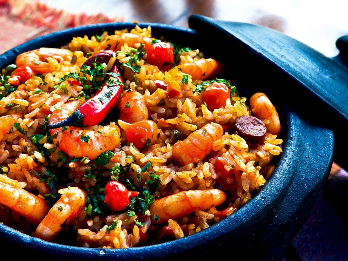 The image shows a delicious dish of paella packed with shrimp, rice, red peppers, and herbs in a black pan with a lid nearby.