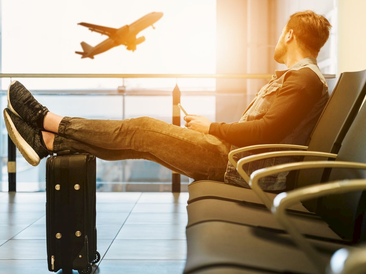 A person sits in an airport lounge with feet resting on luggage, watching a plane take off outside the window.