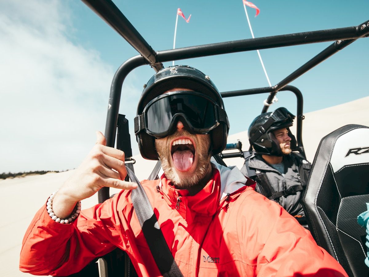 People riding in an off-road vehicle in a sandy area, with one person in front making a hand gesture and sticking their tongue out.