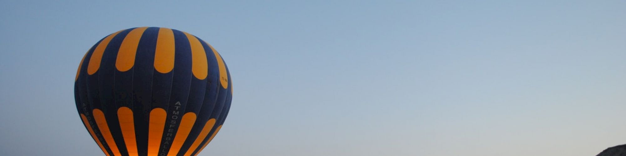 A hot air balloon with blue and orange pattern floats over a rocky landscape at dusk, with a clear sky in the background.