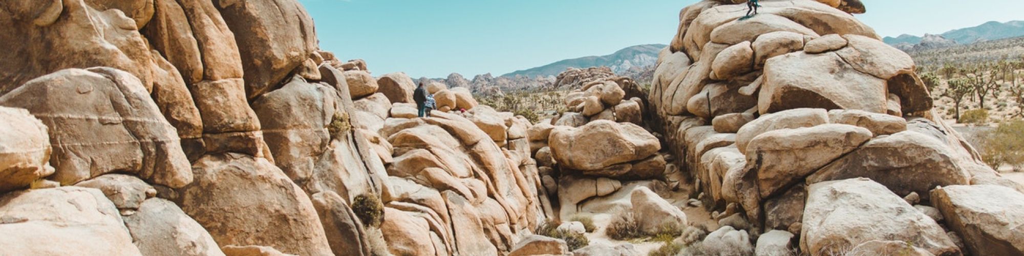 The image shows a rocky desert landscape with large boulders, a clear blue sky, and two people climbing on the rocks.