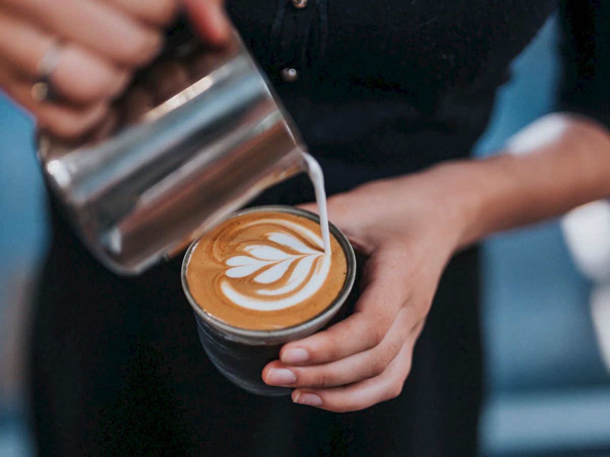 A barista is pouring steamed milk into a cup of coffee to create latte art with a heart-shaped design.