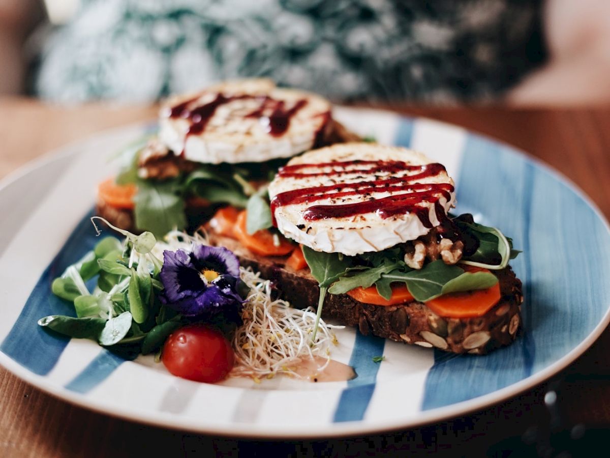 The image shows a plate with a gourmet dish, including greens, grilled cheese, vegetables, and edible flowers, placed on a blue and white plate.