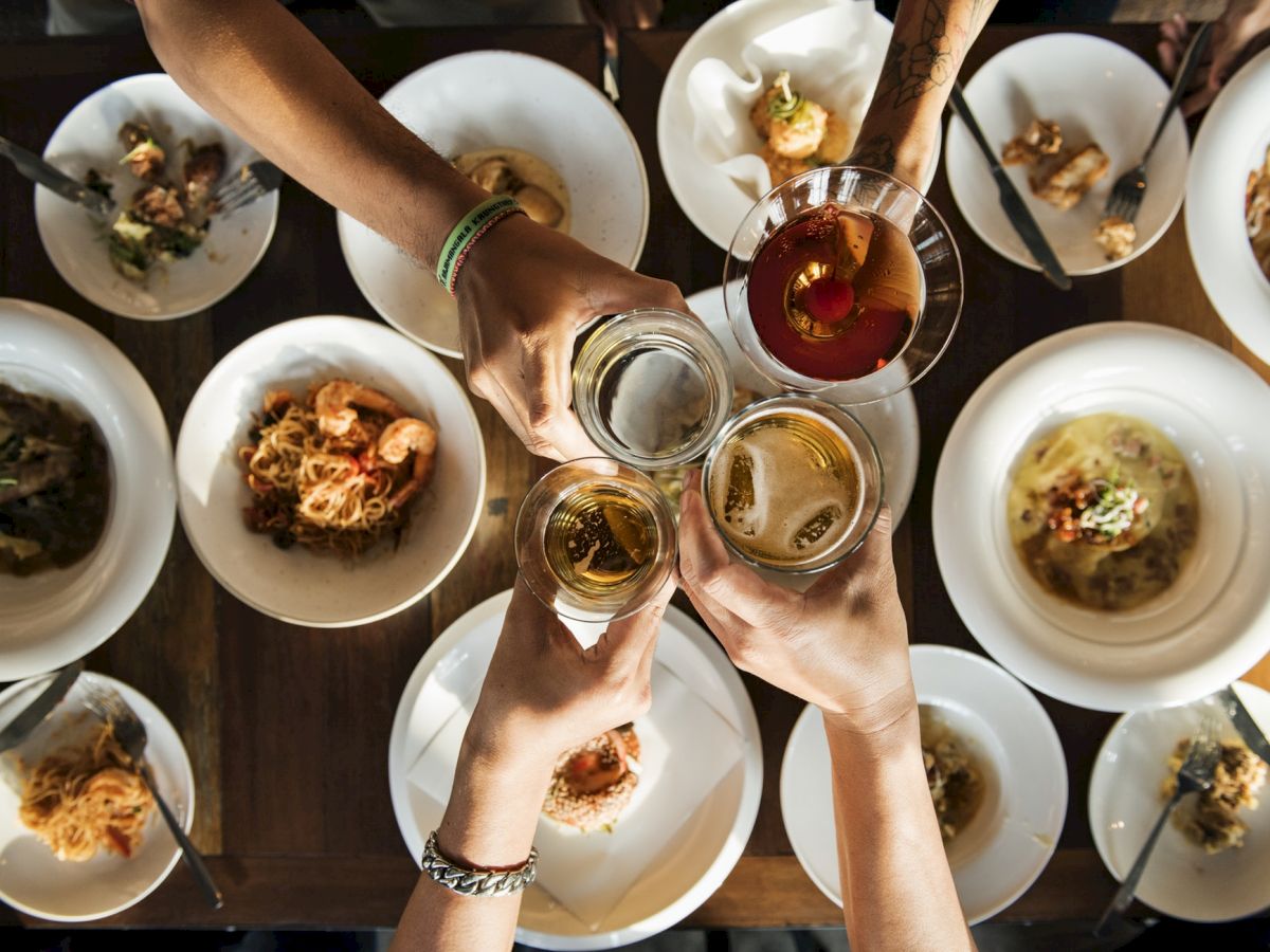 A group of people clinks glasses over a table filled with various dishes, suggesting a meal celebration or gathering.