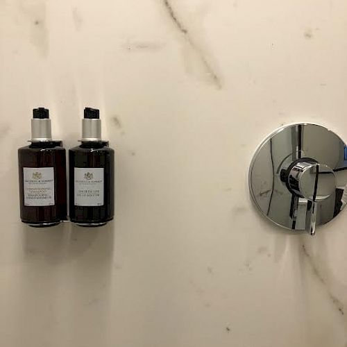 The image shows two mounted bottles of bath products and a chrome shower control on a marble wall.