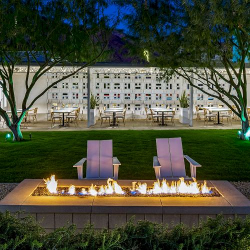 Outdoor seating area with a fire pit, surrounded by chairs and tables under a covered patio, with trees and grass in the foreground.