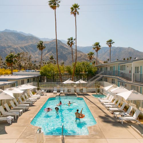 A clear day at a hotel pool, with people swimming, surrounded by lounge chairs and umbrellas, and set against a backdrop of mountains and palm trees.