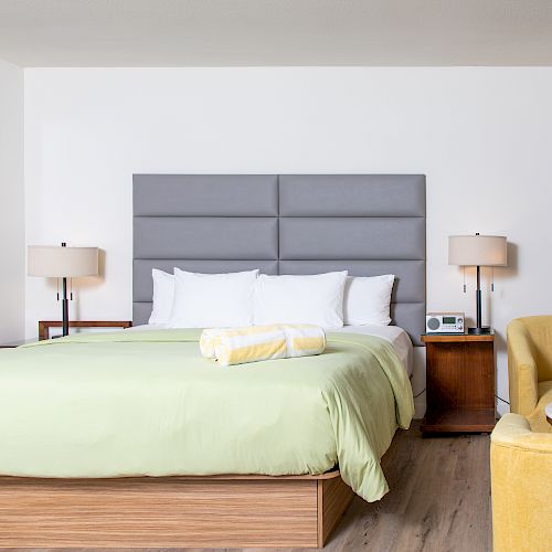 A modern hotel room features a neatly made bed with light green bedding, bedside tables with lamps, a yellow chair, and a framed highway photo.