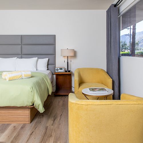 The image shows a bedroom with a bed, yellow armchairs, a nightstand with a lamp, and a window with curtains. The floor is wooden.