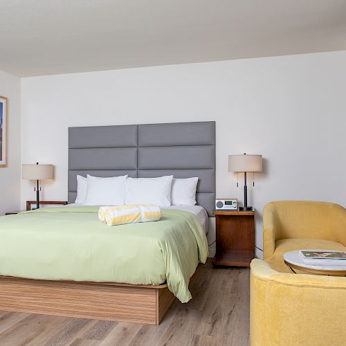 A modern bedroom with a large bed, gray headboard, yellow chair, wooden nightstands with lamps, framed artwork, and a window with gray curtains.