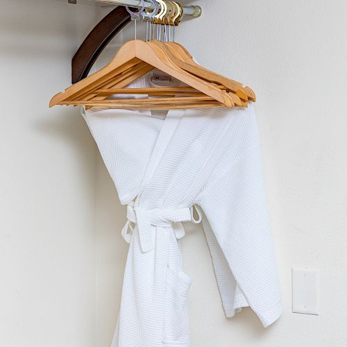 A white robe hanging on wooden hangers in a closet with a shelf above, against a white wall.