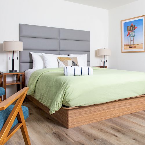 A modern bedroom with a large bed, green bedding, wooden furniture, blue chair, bedside lamps, and a framed artwork on the wall.