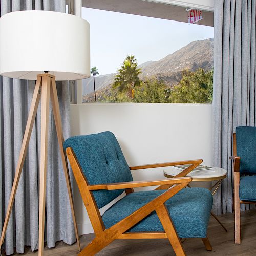 A cozy room with a retro chair, a standing lamp, and a window view of mountains and palm trees, enhanced with grey curtains over a wooden floor.