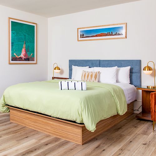 A modern bedroom with a large bed, an orange chair, two framed pictures, bedside tables with lamps, and a floor mirror.