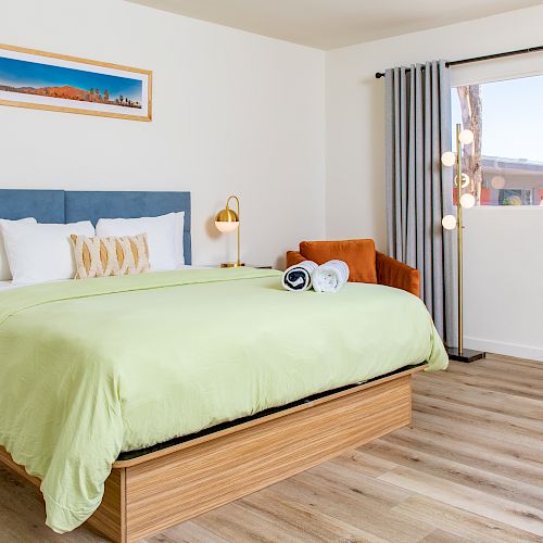 A tidy bedroom with a green duvet, bedside tables, a large window, and a brown chair in the corner. A picture hangs above the bed, ending the sentence.