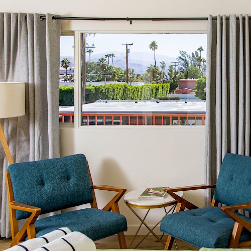 A brightly lit room with two teal armchairs, a small round table, a floor lamp, and gray curtains framing a window overlooking a cityscape.