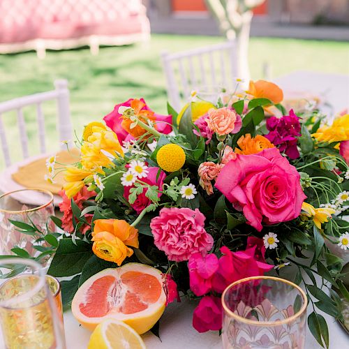 A vibrant table setting with a colorful floral centerpiece, featuring roses and citrus fruits like grapefruit and lemon, surrounded by glassware and cutlery.