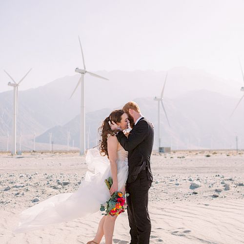 A couple dressed in wedding attire stands in an embrace in a desert landscape with wind turbines in the background.