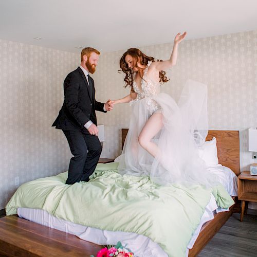 A couple dressed in formal wedding attire is joyfully jumping on a bed, with a bouquet of flowers on the floor beside the bed.