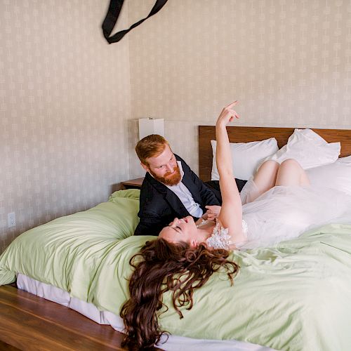 A couple is lying on a bed with a green bedspread. A bouquet of flowers is on the floor in the foreground, and there's a harness on the wall.