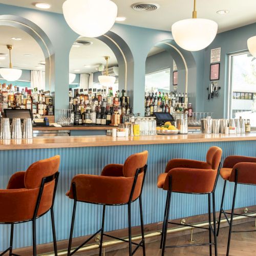 A stylish bar with a blue counter, brown chairs, pendant lights, and a well-stocked selection of bottles is seen in the image.