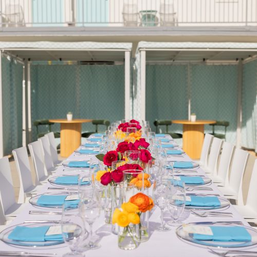 A long table set for an outdoor event with white chairs, blue napkins, clear glasses, and colorful flower centerpieces.